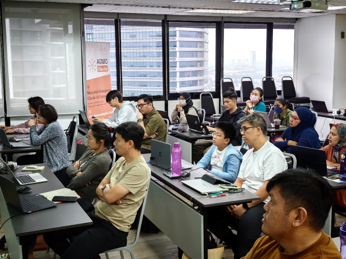 Participants seated at desks with laptops in a training room during the Claude class, with a young boy among the adults