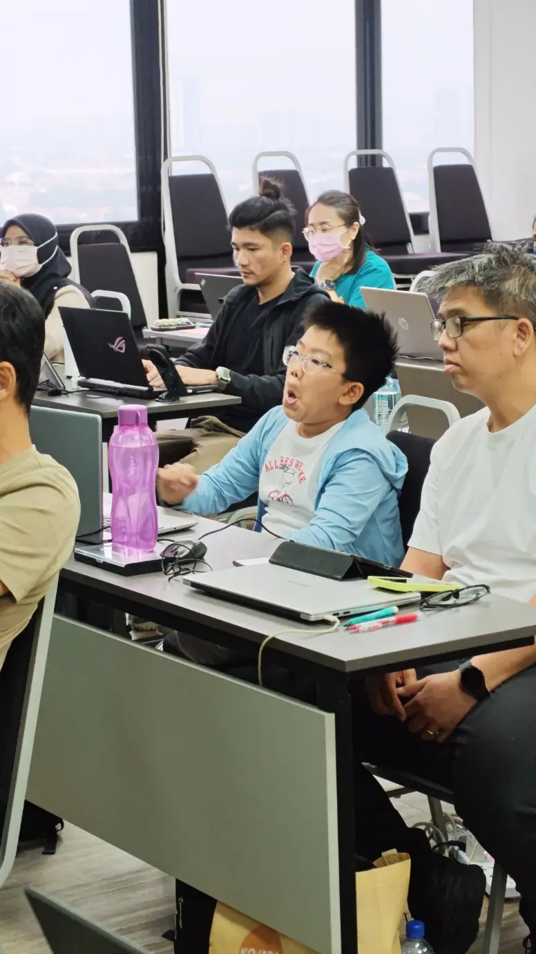 A young boy in a light blue jacket speaking during the class, with other participants seated around him