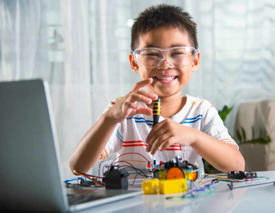 A young student in safety glasses building an electronics project at a laptop