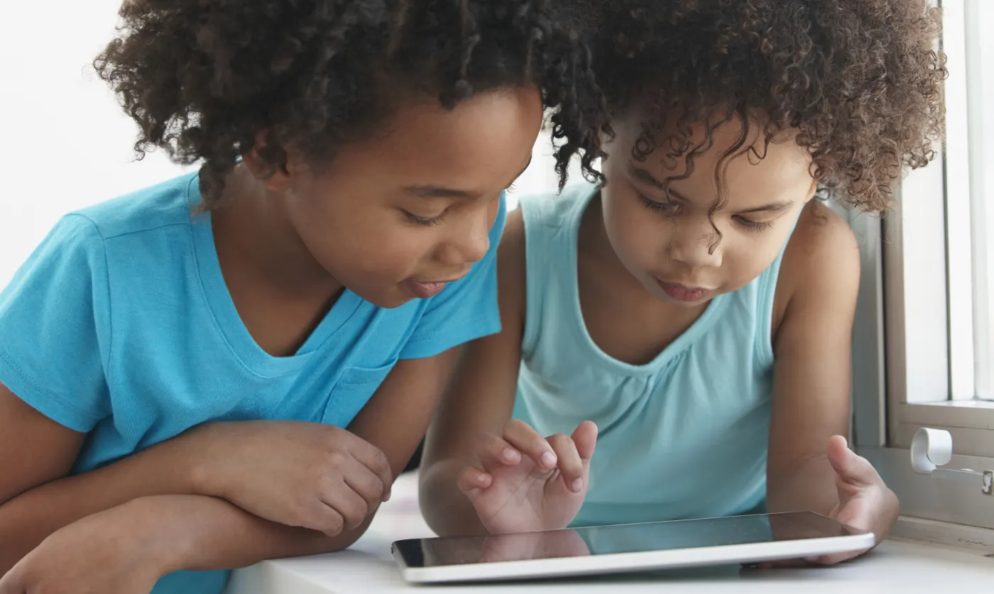 Two children in blue shirts focus intently on a tablet by a window. Bright lighting highlights their concentrated expressions.