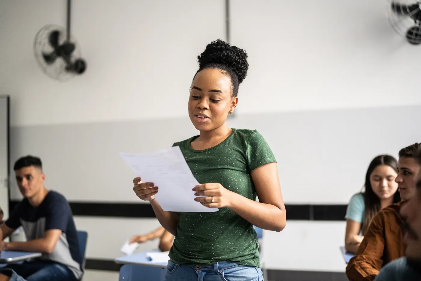 Woman in green shirt reading paper aloud in classroom. Students seated in background. Neutral colors, engaged atmosphere.