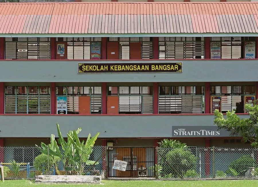 Two-story school building with "Sekolah Kebangsaan Bangsar" sign. Red roof, fenced yard with plants, and visible posters on windows.