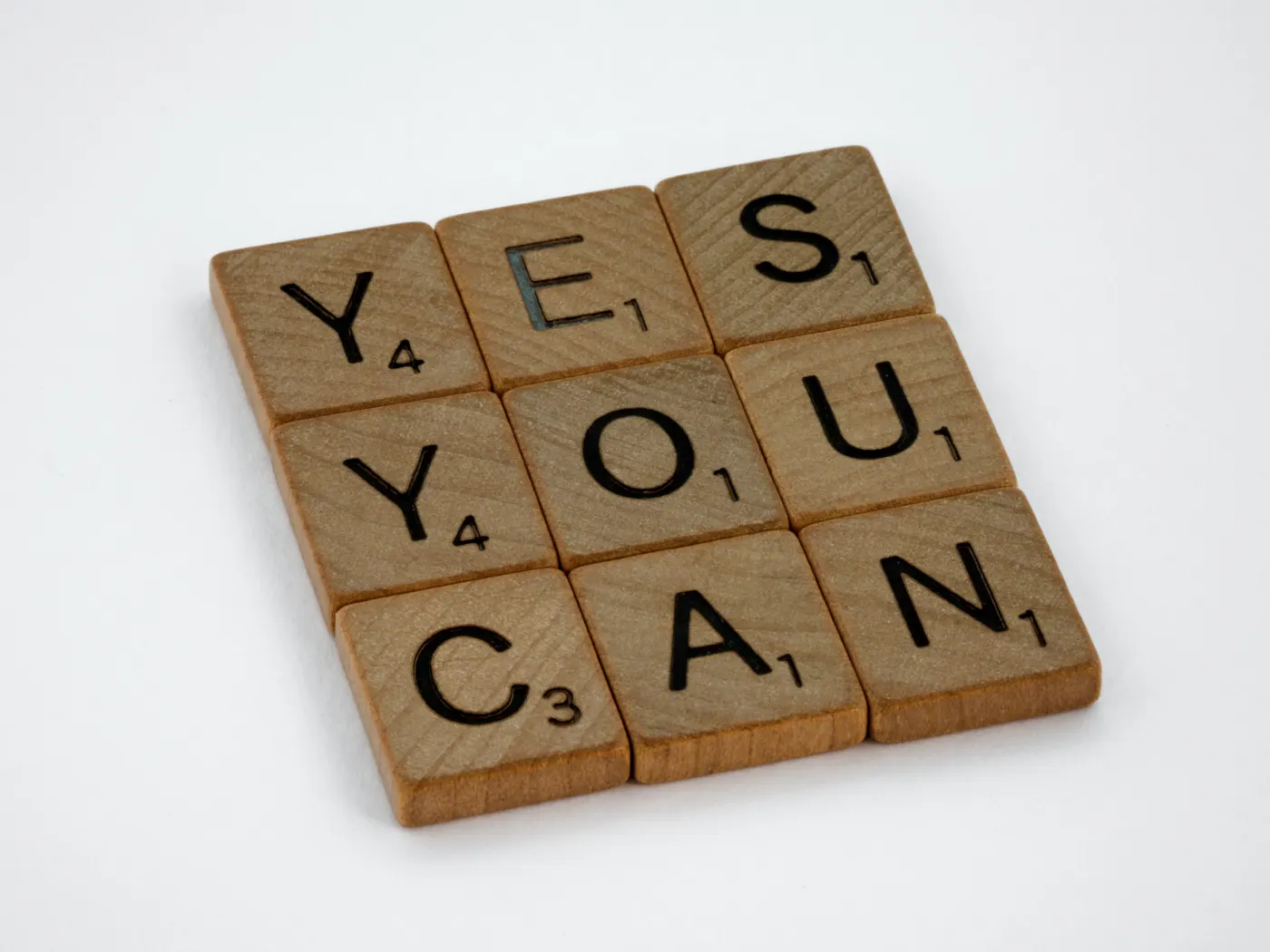 Wooden letter tiles spell "YES YOU CAN" on a white background. Each tile displays a black letter and a small number. Positive mood.