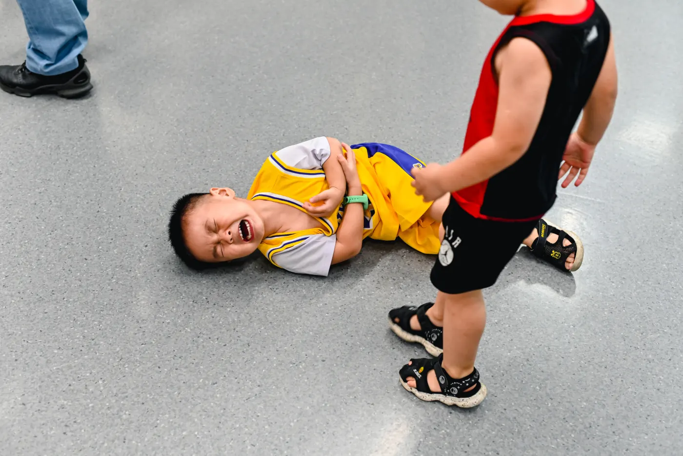 Child in a yellow jersey crying on a gray floor, holding stomach. Another child in red and black stands nearby. Shoes and blue jeans in view.