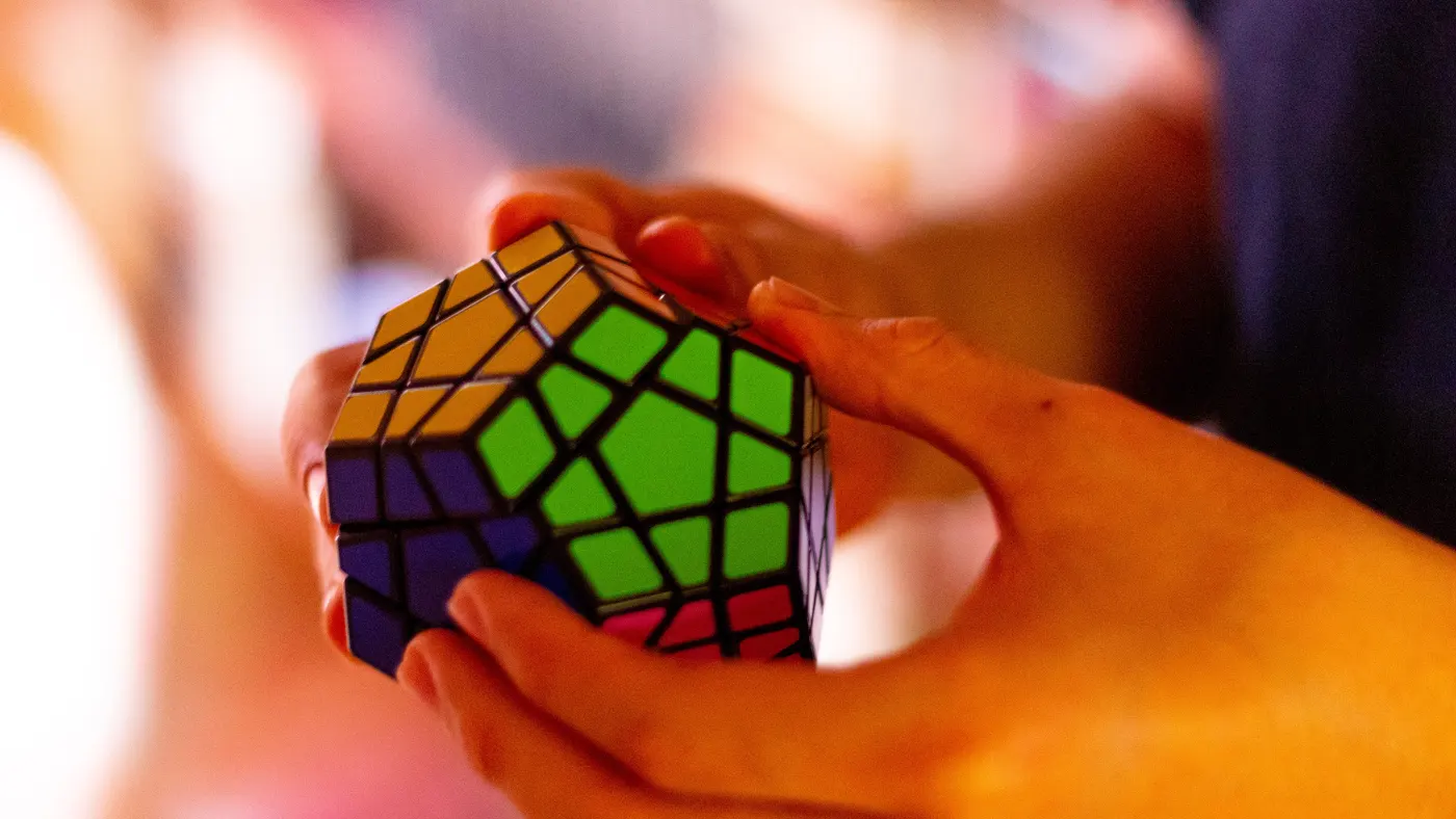 Hands holding a colorful puzzle cube in focus, with a blurred, warm-toned background, conveying concentration and engagement.