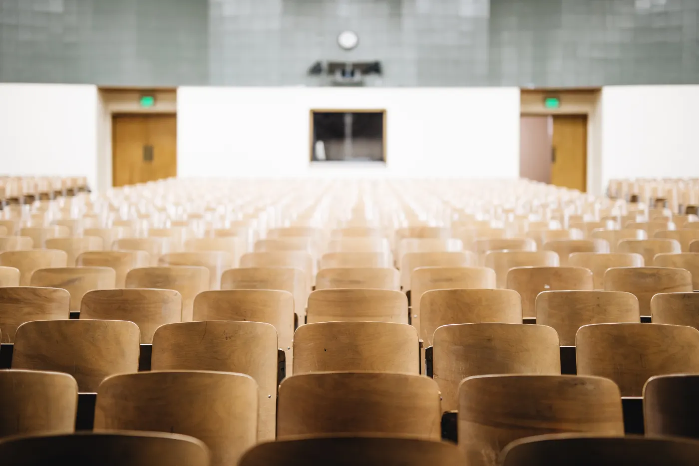 Rows of empty wooden chairs fill a lecture hall. A clock and podium are visible at the front, with doors on both sides. Quiet, academic setting.