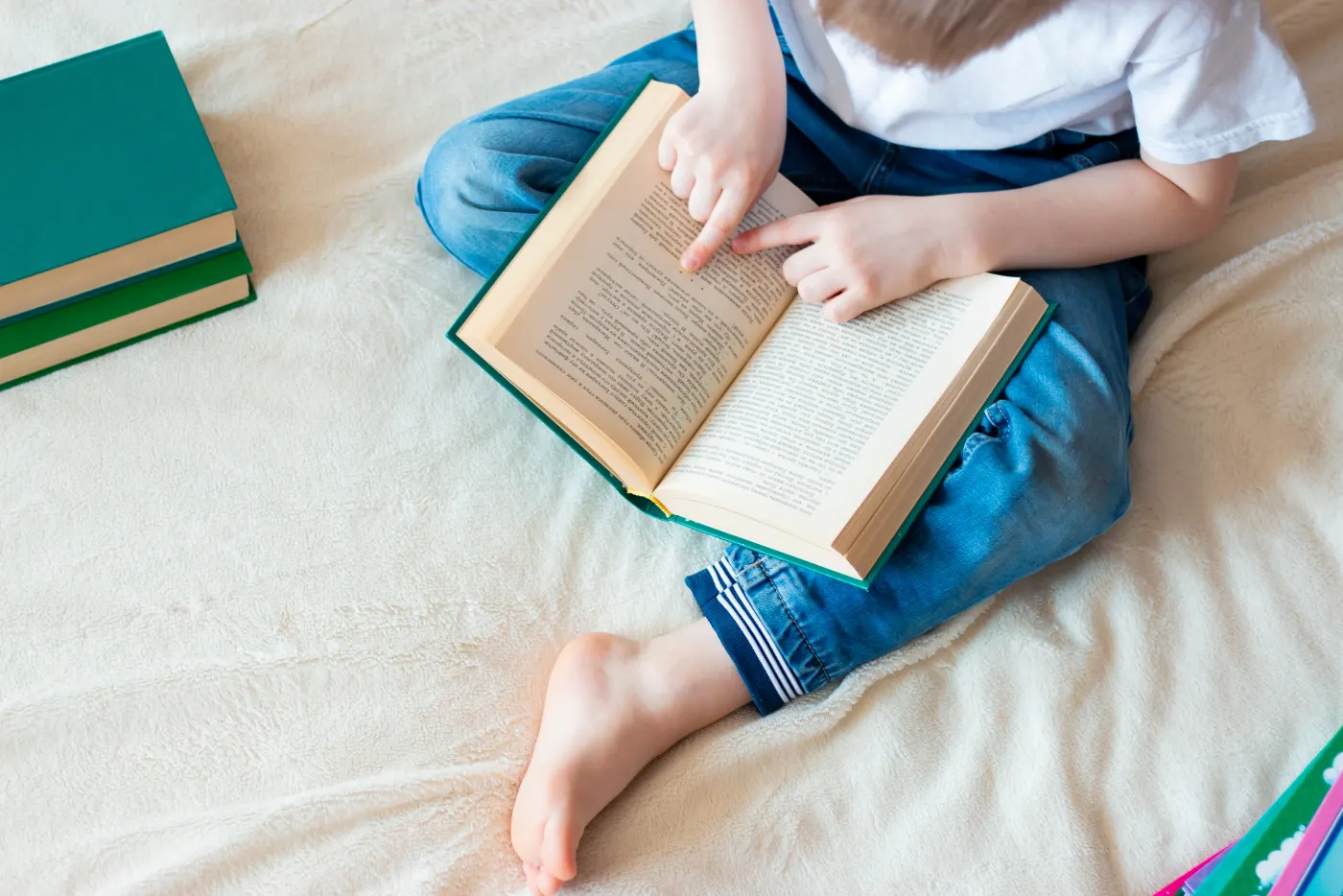 Child in jeans reads a book on a bed, pointing at a page. Nearby are three green books. Cozy setting with a cream blanket.