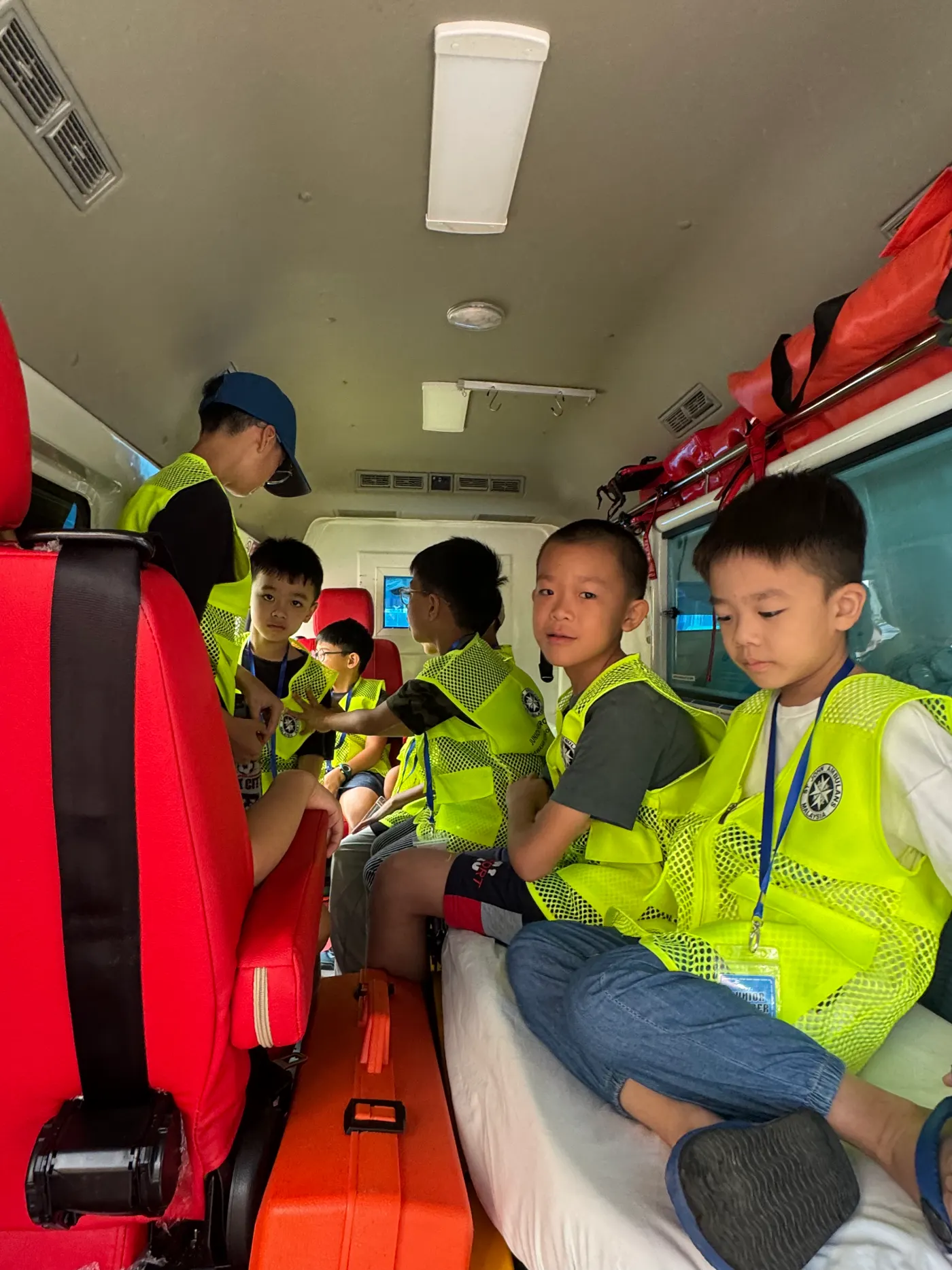 Children in bright yellow vests sit inside an ambulance. Red seats and a first aid kit is visible. The mood is curious and engaged.