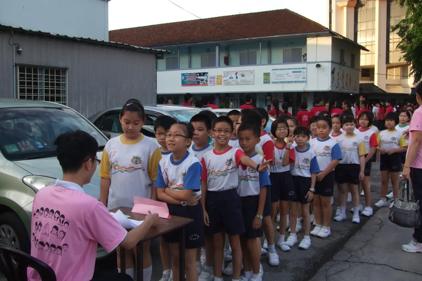 Children in colorful uniforms line up outdoors near a building, awaiting instructions from a seated man. The scene is orderly and calm.