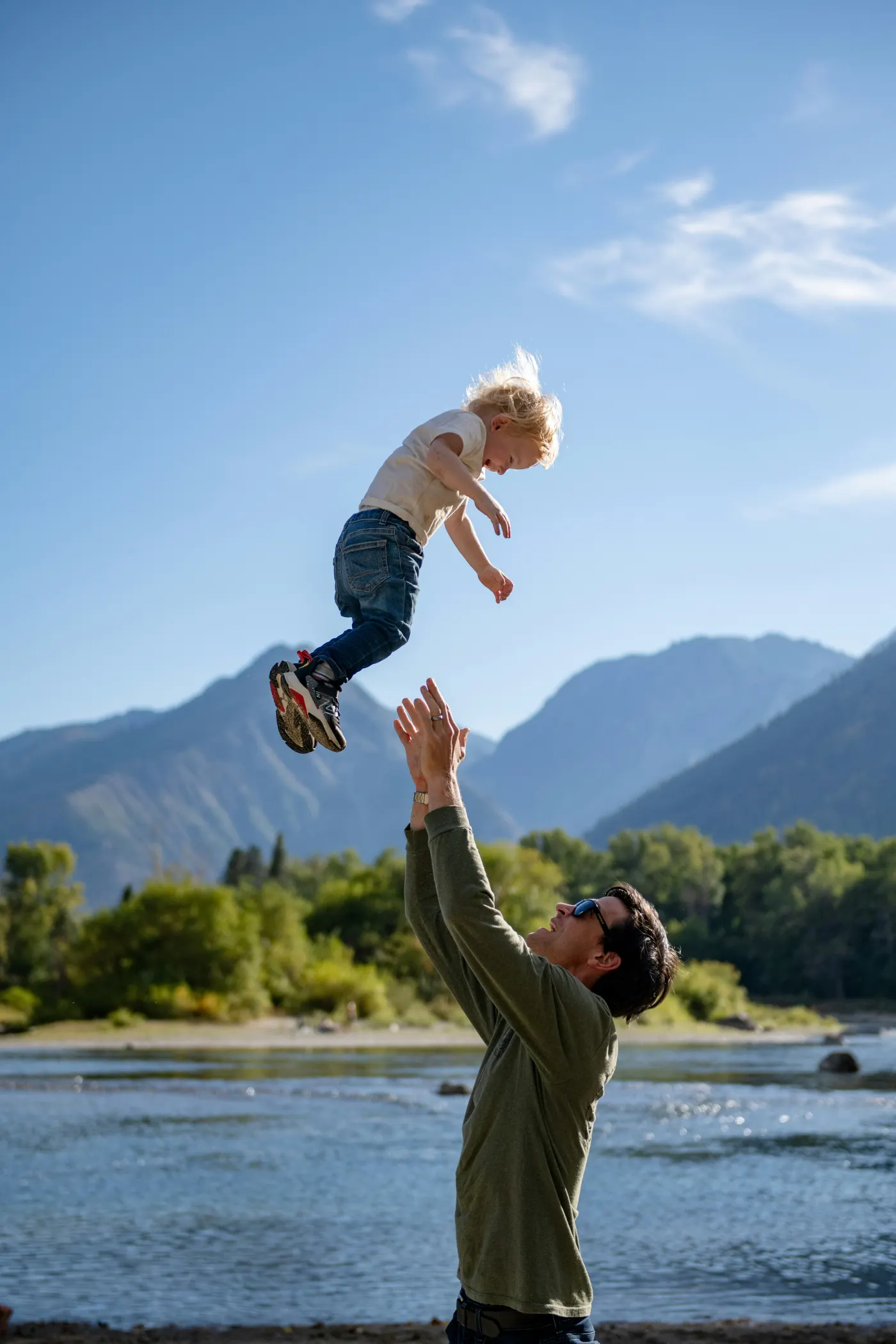 Man tossing smiling child in the air by a river. Mountains and trees in the sunny background. Child in jeans and sneakers, joyful mood.