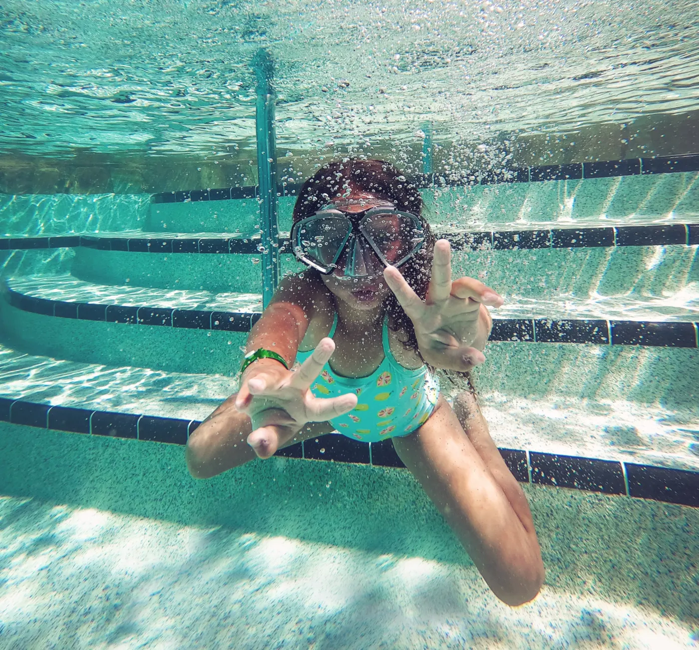 Child underwater in a pool wearing a snorkel. They are playfully posing with fingers extended. Sunlit water creates a bubbly effect.