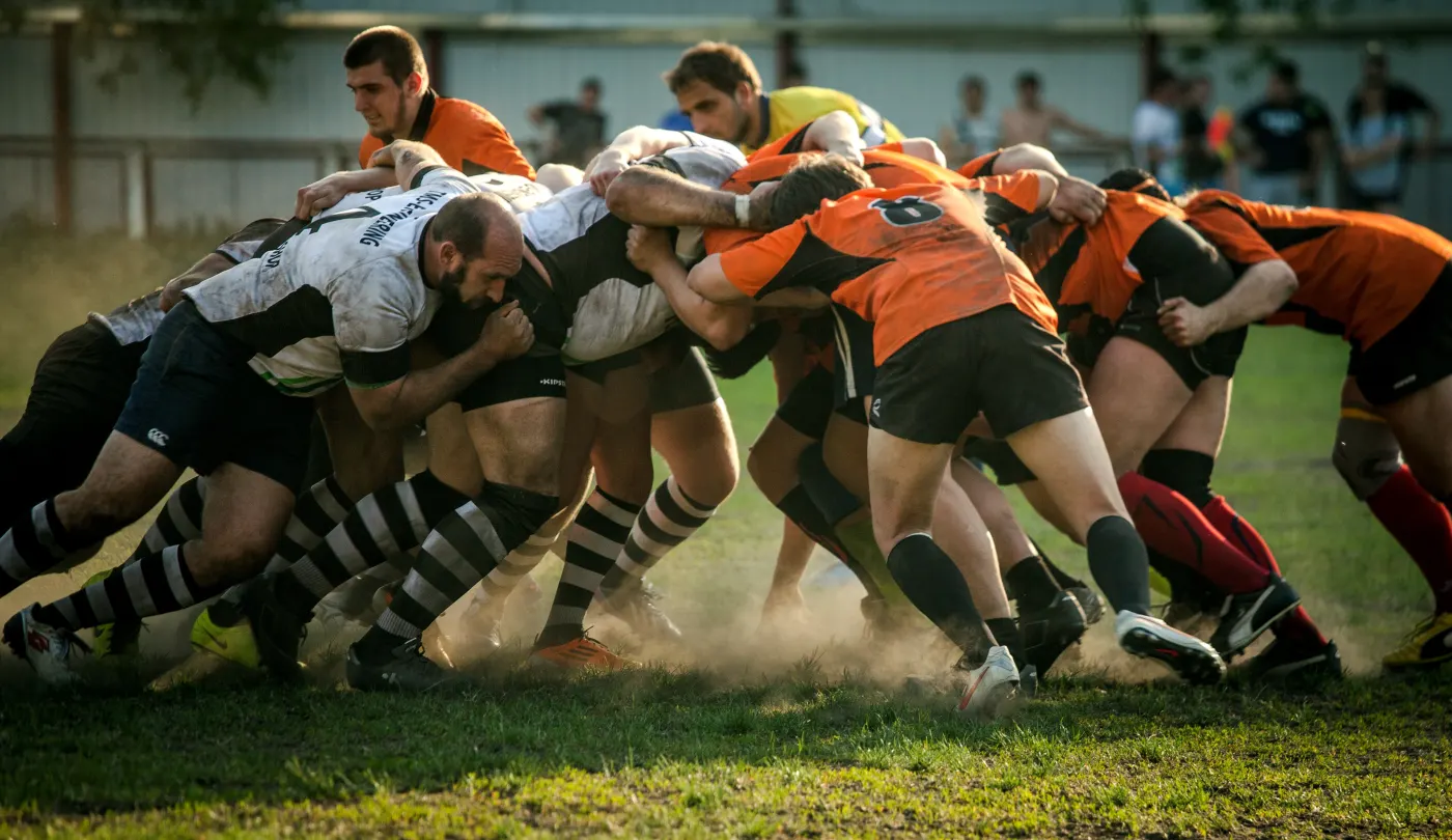 Rugby players in orange and white jerseys engage in a scrum on a grassy field, kicking up dust. Intense focus and teamwork are evident.